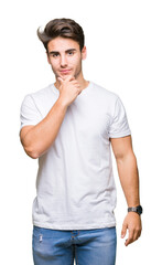 Young handsome man wearing white t-shirt over isolated background looking confident at the camera with smile with crossed arms and hand raised on chin. Thinking positive.