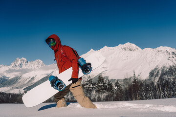snowboarder holding snowboard walking through powder snow against backdrop of sunny mountain...