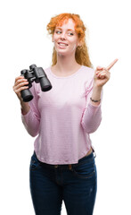 Young redhead woman looking through binoculars very happy pointing with hand and finger to the side