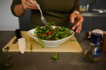 Details on the hands of a woman holding fork and bowl of fresh healthy salad with organic vegetables and greens