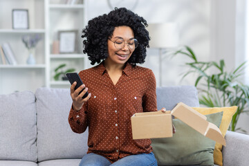 Smiling woman with glasses receiving package at home, happy customer using smartphone on couch