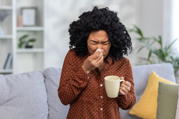 Young woman feeling unwell with cold or flu, sneezing into tissue, holding mug, at home on cozy sofa with cushions - obrazy, fototapety, plakaty