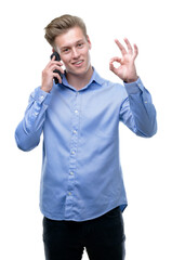 Young handsome blond man using smartphone doing ok sign with fingers, excellent symbol