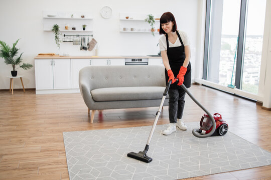 Positive young adult woman in black uniform of cleaning service worker vacuuming carpet in stylish bright studio kitchen. Professional female cleaner in apron actively cleans modern apartment.