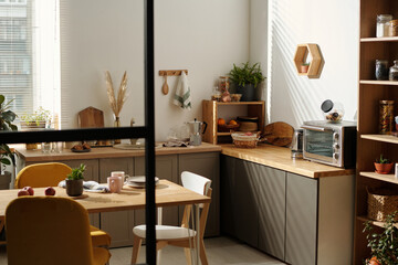 Corner of modern kitchen with counter standing along wall, wooden table with kitchenware, green...