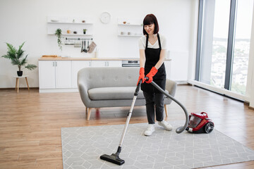 Smiling Caucasian female cleaning service worker vacuums rug in living room kitchen with modern...