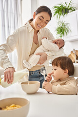 cheerful woman pouring milk into corn flakes for her toddler son and holding her newborn baby