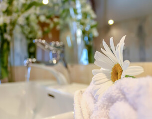 Photograph of a daisy with white petals on white towels, with a background blurred with plants and a stainless steel faucet from which a stream of water comes out. Wellness and spa concept