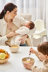 Obraz na płótnie Canvas loving good looking mother having breakfast with her toddler son and holding her newborn baby