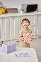 Fototapeta na wymiar cute toddler boy in homewear holding flower bouquet near table with gift and postcard on it