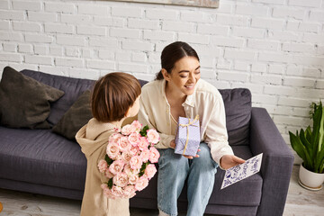 cute toddler boy giving his jolly mom present and postcard with flower bouquet on Mothers day