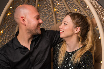 Portrait of an elegant couple. A young woman and a man are sitting in a hanging chair decorated with glowing lights. They hug and look at each other tenderly