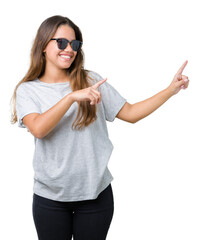 Young beautiful brunette woman wearing sunglasses over isolated background smiling and looking at the camera pointing with two hands and fingers to the side.