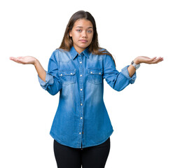 Young beautiful brunette woman wearing blue denim shirt over isolated background clueless and confused expression with arms and hands raised. Doubt concept.