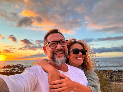 Happy adult couple in love taking selfie picture with romantic wonderful sunset on the ocean in background. Young mature people man and woman enjoying travel destination in summer holiday vacation