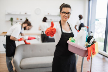 Portrait of young African American woman professional cleaning worker holding a bucket for washing with detergents showing thumb up, on bright kitchen studio background, copy space.