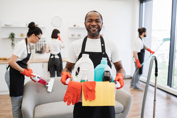 Portrait of young African American man professional cleaning worker holding a bucket for washing with detergents on bright kitchen studio background, copy space. - obrazy, fototapety, plakaty