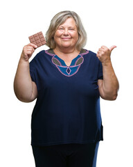 Obraz premium Senior plus size caucasian woman eating chocolate bar over isolated background pointing and showing with thumb up to the side with happy face smiling