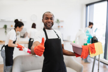 Portrait of young African American man professional cleaning worker holding a bucket for washing with detergents showing thumb up, on bright kitchen studio background, copy space.
