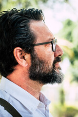 Fototapeta premium Side view of portrait adult mature man with beard and glasses looking and thinking - green park outdoor background - concept of serious people worried about problems