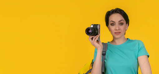 smiling woman photographer, a travel blogger with backpack taking photos with camera,  studio shot,  yellow background. - obrazy, fototapety, plakaty