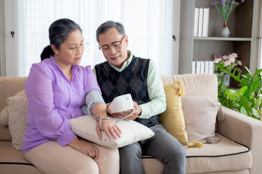 Asian senior couple sitting on sofa checking blood pressure with pressure gauge in living room at home, elderly man and woman sitting on couch checkup health and pressure, medical of patient.
