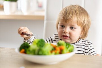 Cute little girl is eating broccoli and carrot vegetables with a fork