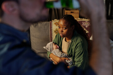 Fototapeta na wymiar Focus on young upset and annoyed African American woman with baby on hands looking at her husband drinking beer from bottle