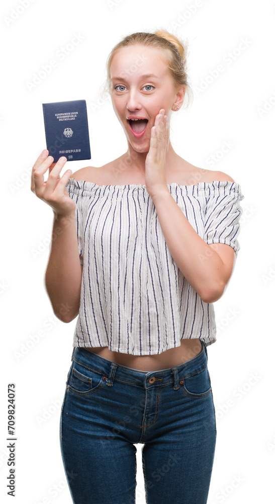 Sticker blonde teenager woman holding passport of germany scared in shock with a surprise face, afraid and e