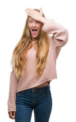 Blonde teenager woman wearing pink sweater surprised with hand on head for mistake, remember error. Forgot, bad memory concept.