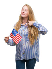 Blonde teenager woman holding flag of United States of America with surprise face pointing finger to himself