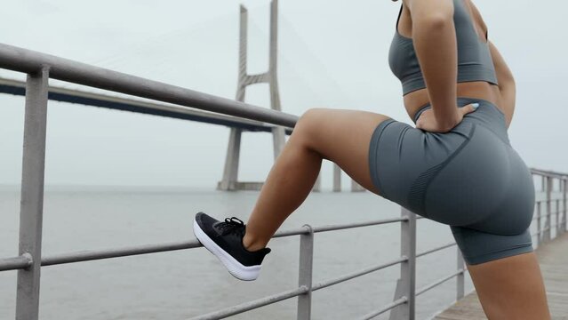 Unseen sporty woman in tight activewear stretching her legs, preparing for a run on a bridge by the riverside