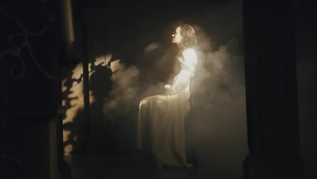 A young woman in a white long dress sits on a chair in a dark Gothic church. Bright sun rays and contrasting shadows. Prayer. Meditation and expression. Smoke. Fashion and Cinematic concept