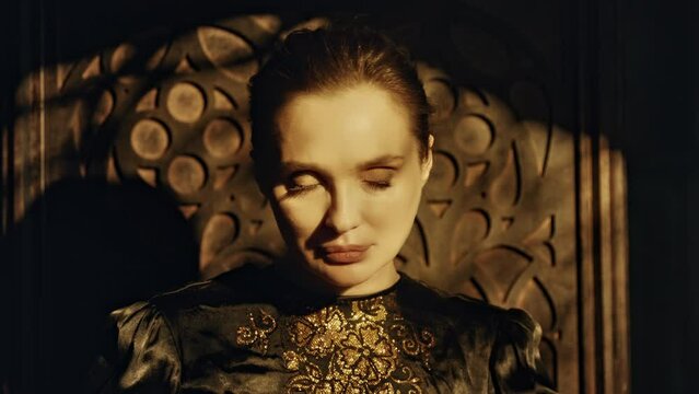 Portrait of a young woman in a black long dress sits in a wooden carved throne in a dark Gothic church. Looks at the camera and closes his eyes. Meditation and expression. Fashion and Cinematic 