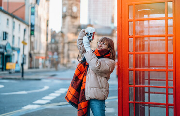 Outdoor portrait of young happy woman using a vintage camera   against red phone box in English city