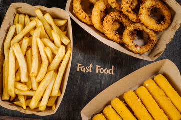 Set of takeaway paper boxes with different fast food, french fries, nuggets, onion rings and different sauces on wooden background. View from above