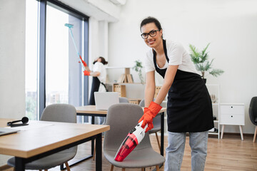 Happy smiling multicultural employees of company cleaning in bright, spacious modern office. Multiethnic young female cleaner vacuuming chair with portable cordless vacuum cleaner, looking at camera.