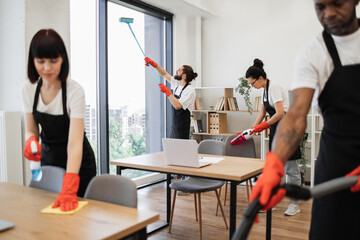 Professional team of people of cleaning service in black aprons and red gloves cleans tables, floor, and panoramic windows of spacious studio . Multinational group cleans bright office.