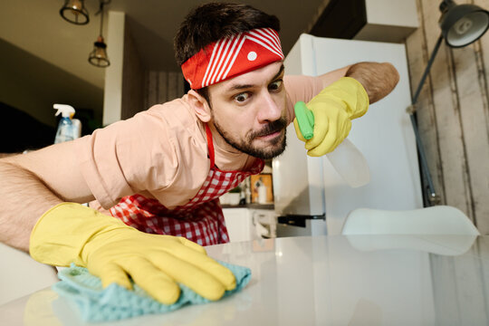 Guy in apron, headband and rubber gloves wiping table with duster and spraying detergent while carrying out domestic chores