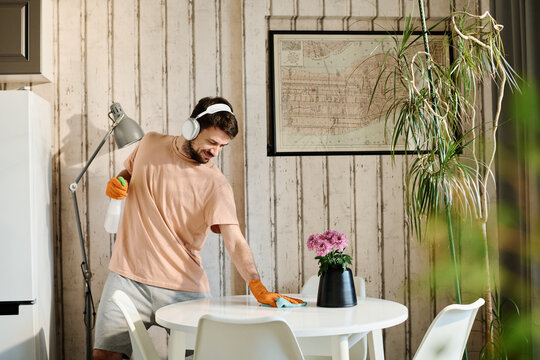 Young man in headphones, casualwear and rubber gloves wiping kitchen table and spraying detergent while carrying out domestic chores