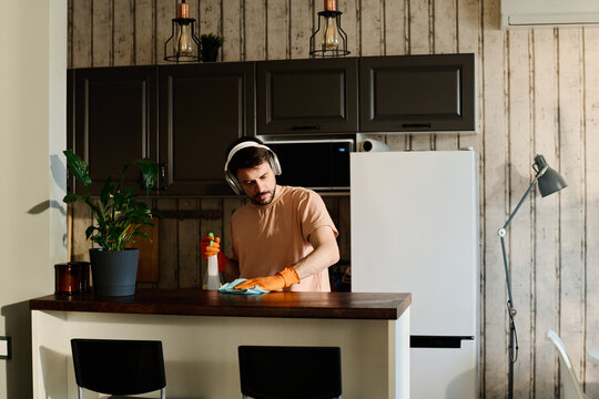 Young man listening to music in headphones and wiping kitchen counter with duster and detergent while carrying out domestic chores