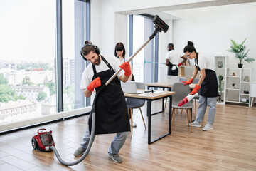 Professional cleaner Caucasian man having fun holding vacuum cleaner like guitar imitating playing at concert . Happy multinational team of people in black aprons cleaning modern office.