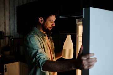 Young man with plastic bottle of fresh milk opening door of refrigerator and looking at food...