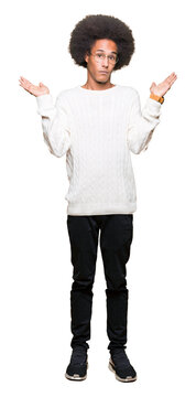 Young african american man with afro hair wearing glasses clueless and confused expression with arms and hands raised. Doubt concept.