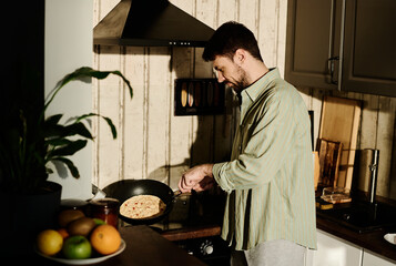 Side view of young man in grey cotton shirt holding hot frying pan while standing by electric stove and cooking omelet for breakfast