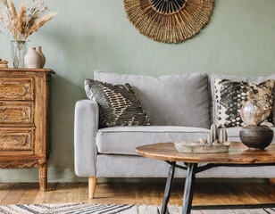 Chic boho living room interior featuring a design gray sofa, wooden coffee table, commode, and elegant personal accessories, blending style and comfort seamlessly.