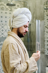 Side view of young bearded man in bathrobe standing in bathroom and looking at Y-shaped ball roller...
