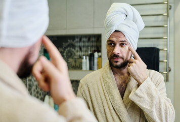 Young bearded man with towel on head putting moisturizing patches on under eye area while having beauty procedure after morning shower