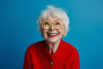 Happy woman with gray hair, 70 years old, wearing a red cardigan and round yellow glasses - obrazy, fototapety, plakaty