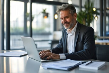 Fototapeta premium Happy mid aged professional business man company executive ceo manager lawyer sitting at desk in office using laptop working on plan overview checking, Generative AI